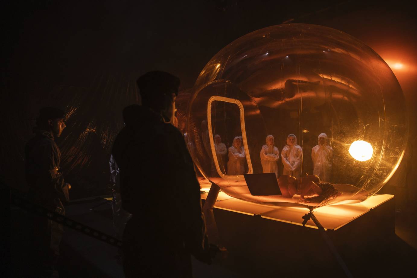 In the foreground, a woman crouches; in the background, three people in protective gear look at her through a bubble